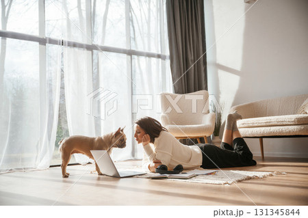 In sunlight. Lying down on floor with laptop. Looking at pet. Young woman is with her pug dog at home In sunlight. Lying down on floor with laptop. Looking at pet. Young woman is with her pug dog at home 131345844