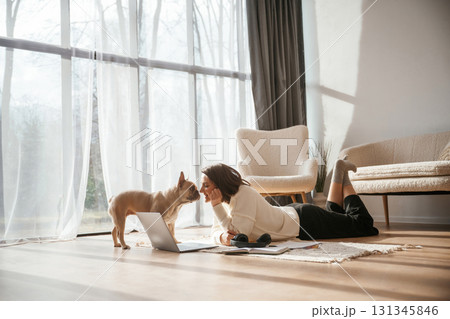 In sunlight. Lying down on floor with laptop. Looking at pet. Young woman is with her pug dog at home 131345846