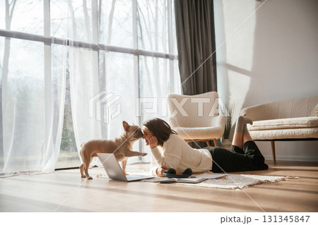 In sunlight. Lying down on floor with laptop. Looking at pet. Young woman is with her pug dog at home In sunlight. Lying down on floor with laptop. Looking at pet. Young woman is with her pug dog at home 131345847