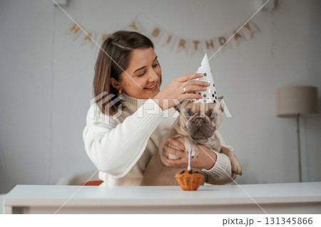 Happy birthday. Pet in hat and with cake. Young woman is with her pug dog at home Happy birthday. Pet in hat and with cake. Young woman is with her pug dog at home 131345866