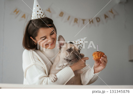 Birthday of the pet. In festive hats and with cake. Young woman is with her pug dog at home 131345876