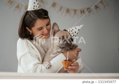 Birthday of the pet. In festive hats and with cake. Young woman is with her pug dog at home 131345877