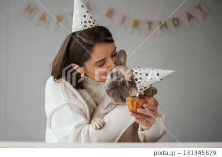 Happy owner is celebrating birthday of pet. In hat and with cake. Young woman is with her pug dog at home 131345879