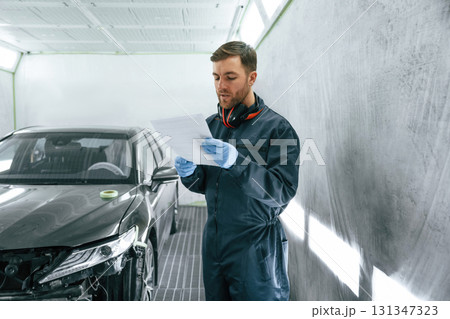Reading information. Car painter is standing in the garage with documents in hands 131347323