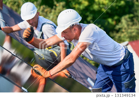 Workers building solar panel system on rooftop of house. Two men installers in helmets and gloves installing photovoltaic solar module outdoors. Alternative and renewable energy generation concept. 131349134