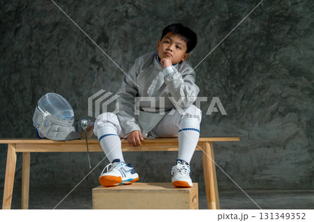 Asian boy with fencing uniform put epee and mask beside and sit on wooden bench to thinking about the competition during relax from practice. Asian boy with fencing uniform put epee and mask beside and sit on wooden bench to thinking about the competition during relax from practice. 131349352