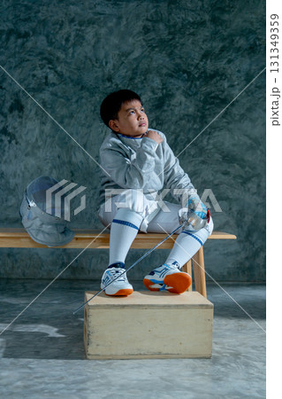 Vertical image of boy wear fencing sport uniform also hold epee and sit on wooden bench with look up to his left side. Vertical image of boy wear fencing sport uniform also hold epee and sit on wooden bench with look up to his left side. 131349359