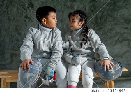 Asian brother and sister with fencing sport uniform sit beside on wooden bench and look to each other and smile also relax after practice for competition. 131349424