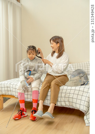 Asian mother sit on bed next to her daughter and set hair of her child during practice fencing sport in bedroom with soft morning light and happiness. Asian mother sit on bed next to her daughter and set hair of her child during practice fencing sport in bedroom with soft morning light and happiness. 131349428