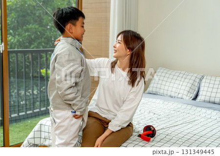 Close up Asian mother sit on bed and touch shoulder of her son to encourage during practice fencing sport in bedroom with soft morning light and happiness. Close up Asian mother sit on bed and touch shoulder of her son to encourage during practice fencing sport in bedroom with soft morning light and happiness. 131349445