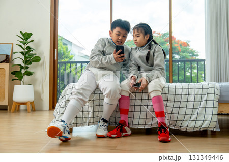 Frong view of boy and girl with uniform of fencing sport sit on bed together and discuss about the practice using mobile phone. Frong view of boy and girl with uniform of fencing sport sit on bed together and discuss about the practice using mobile phone. 131349446