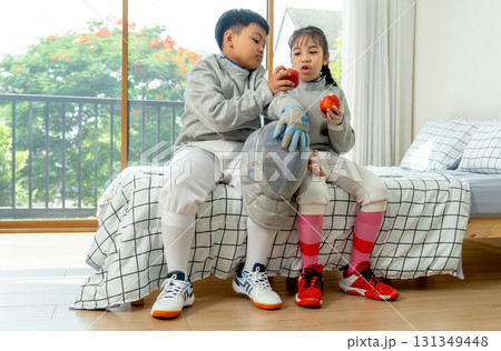 Boy and girl with uniform of fencing sport sit on bed together during enjoy eating apple in bedroom during relax after practice. 131349448