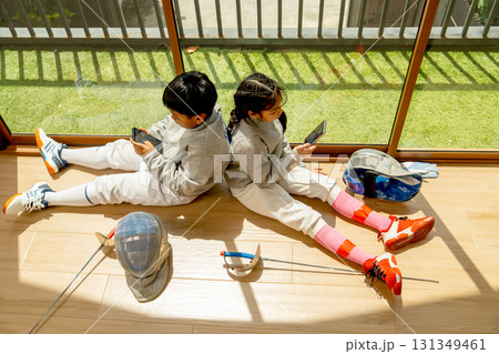 Asian boy and girl with fencing sport uniform enjoy to play mobile phone and sit on the floor in front of glass door with warm light after practice for competition. 131349461