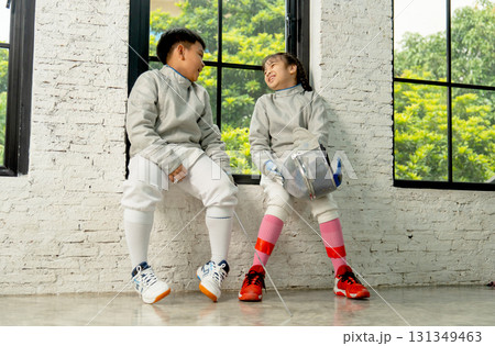 Brother and sister with fencing sport uniform sit near the glass windows and talk together after relax from practicing. Brother and sister with fencing sport uniform sit near the glass windows and talk together after relax from practicing. 131349463