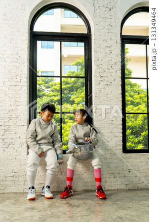 Vertical image of brother and sister with fencing sport uniform sit near the glass windows and talk together after relax from practicing. Vertical image of brother and sister with fencing sport uniform sit near the glass windows and talk together after relax from practicing. 131349464