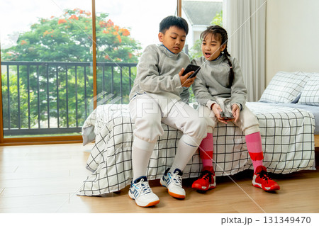 Boy and girl with uniform of fencing sport sit on bed together and discuss about the practice using mobile phone. Boy and girl with uniform of fencing sport sit on bed together and discuss about the practice using mobile phone. 131349470