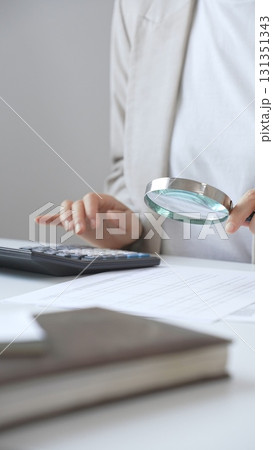 Businesswoman carefully reviewing financial reports using magnifying glass and calculator, close up vertical view. Audit and taxes in business Businesswoman carefully reviewing financial reports using magnifying glass and calculator, close up vertical view. Audit and taxes in business 131351343