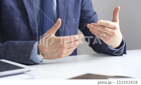 Professional businessman gesturing while discussing strategy, seated at white desk with tablet, eyeglasses, and wristwatch reflecting on sleek workspace surface. Business people concept 131351419