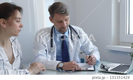 Male doctor writing notes on a clipboard while attentively listening to a female patient during a medical consultation in a hospital office, fostering communication and understanding Male doctor writing notes on a clipboard while attentively listening to a female patient during a medical consultation in a hospital office, fostering communication and understanding 131351429