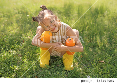 A cute, cheerful girl in rubber boots hugs two large yellow squash. The harvest season in the vegetable garden A cute, cheerful girl in rubber boots hugs two large yellow squash. The harvest season in the vegetable garden 131351563