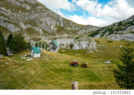 Two small houses and a red tractor standing on a grassy hill among mountains, rural farming landscape in daylight 131351681
