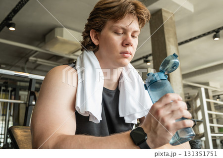 Young sporty man sitting, relax and holding water bottle and towel at gym. Moment of relaxation and refreshment after workout. Young sporty man sitting, relax and holding water bottle and towel at gym. Moment of relaxation and refreshment after workout. 131351751