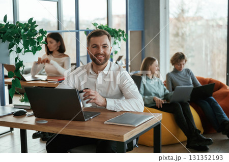 Cheerful friendly man is sitting by the table with laptop. People are working in the office with bean bags chairs in it 131352193