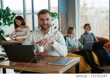 Man is smiling, sitting by the table with laptop. People are working in the office with bean bags chairs in it 131352197