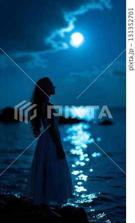 Woman in white dress stands by the ocean under a bright moonlight at night, creating a serene atmosphere 131352701