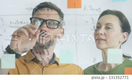 Professional businesspeople collaborating, reviewing data on transparent whiteboard, developing strategic corporate solutions in modern collaborative workspace. Business people concept 131352771