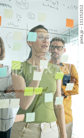 Professional businesswoman wearing glasses writing on glass board, marking strategic plan with sticky notes while colleagues brainstorming in background Professional businesswoman wearing glasses writing on glass board, marking strategic plan with sticky notes while colleagues brainstorming in background 131352812