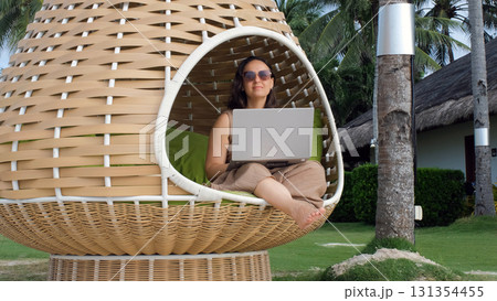 Female freelancer works on her laptop from a woven chair surrounded by palm trees and a beautiful beach view blending work and relaxation Female freelancer works on her laptop from a woven chair surrounded by palm trees and a beautiful beach view blending work and relaxation 131354455