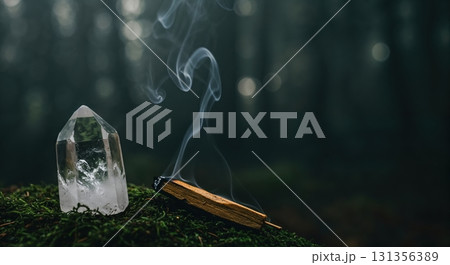 A clear quartz crystal and a smoldering Palo Santo stick producing ethereal smoke rest on vibrant green moss within a serene, blurred forest setting. 131356389