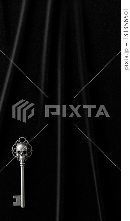 An elegant silver skeleton key with a skull motif on its bow rests on a dark, textured fabric background, creating a mysterious and gothic atmosphere. 131356501