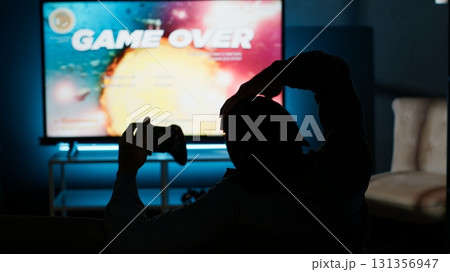 Male gamer getting frustrated with his failure and lost gaming competition, angry young man losing video games challenge online with friends. Modern smart tv setup for game night at home. Camera B. 131356947