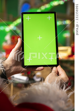 Close up of Santa using green screen tablet in festive workshop to check messages from kids worldwide. Father Christmas reads holidays wishes sent by children via email using chroma key device during 131356991