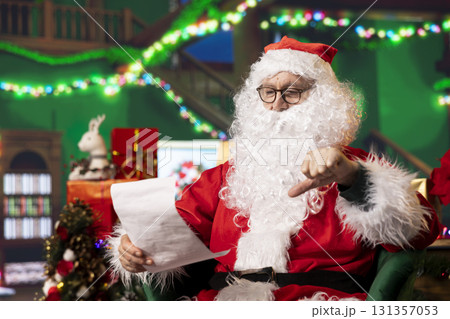 Santa Claus gives disapproving thumbs down sign while reading Christmas letters, answering requests. Upset man in xmas suit looks at wish lists to fulfill kids hopes, doing negative hand gesture 131357053