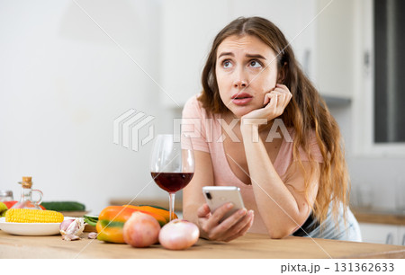 Upset woman with glass of wine in kitchen reading messages on mobile phone screen Upset woman with glass of wine in kitchen reading messages on mobile phone screen 131362633