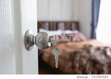 Close-Up View of a Door Knob with a Key Hanging, Soft Focus on a Cozy Bedroom Interior with Brown Bedding and Natural Light Streaming In Close-Up View of a Door Knob with a Key Hanging, Soft Focus on a Cozy Bedroom Interior with Brown Bedding and Natural Light Streaming In 131365383