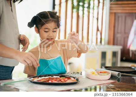 Young girl preparing homemade pizza in a kitchen with parents, focusing on creativity and joy of cooking together with fresh ingredients and teamwork 131366364