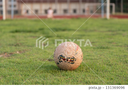 Worn Soccer Ball on Grass Field in Soft Evening Light with Blurred Background of Soccer Goal and Children Playing in Distance 131369096