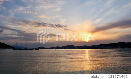 Sun dips below the hills, painting the sky with warm hues as it reflects off the gentle sea, with sailboats silhouetted on the tranquil water. Sun dips below the hills, painting the sky with warm hues as it reflects off the gentle sea, with sailboats silhouetted on the tranquil water. 131371461