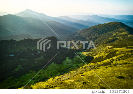 Aerial view of rugged mountain range with lush green slopes at sunset. Landscape showcases beauty of untouched nature with distant peaks and valleys bathed in soft sunlight. Carpathians, Chornogora. 131371802