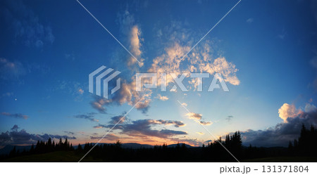 Serene evening sky with hues of blue and soft pink clouds as sun sets. Horizon glows gently, casting light over silhouetted pine trees and rolling hills, creating peaceful and expansive landscape. 131371804