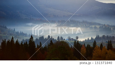 Misty autumn morning in mountain valley. Layers of hills bathed in soft light, with patches of sunlight breaking through fog, illuminating autumn foliage. Interplay of light and shadow. 131371840