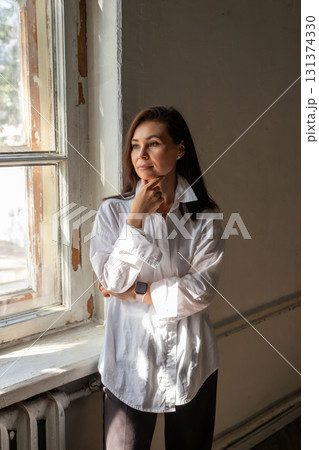 Woman in a white shirt standing thoughtfully near a window in a bright room during daytime. A business portrait of woman in creative profession. Architect, designer, artist, photographer, doctor 131374330