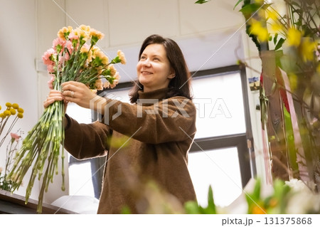 woman florist is skillfully arranging pink flowers into a bouquet amidst a cozy flower shop filled with various blossoms. Sunlight streams through the window, enhancing the inviting atmosphere. 131375868