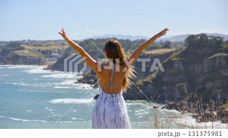 Young woman raising arms enjoying freedom by ocean cliff 131375981