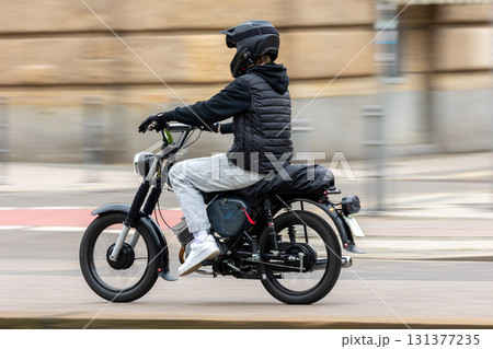 Young motorcyclist in casual clothing and helmet rides black motorbike along urban street. Dynamic shot with motion blur emphasizes speed, energy and the urban lifestyle of two-wheeled transport Young motorcyclist in casual clothing and helmet rides black motorbike along urban street. Dynamic shot with motion blur emphasizes speed, energy and the urban lifestyle of two-wheeled transport 131377235