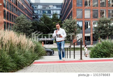 Businessman walking from office building, holding tablet and reading something. 131378083